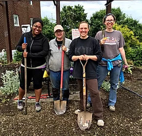 Four people smiling and holding garden tools in a garden.